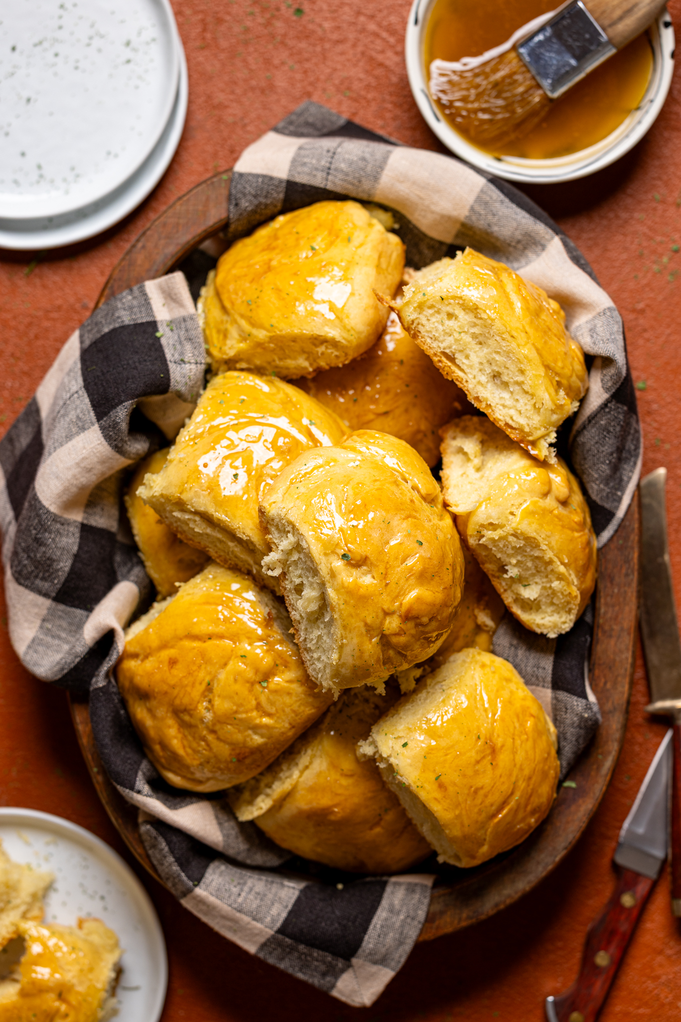 Baked dinner rolls in a basket with checkered cloth, plates, and honey-butter mixture. 