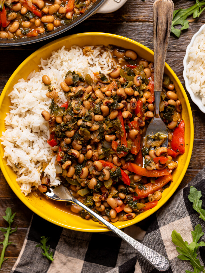 Up close shot of cooked black eyed peas in a yellow bowl with rice and utensils on a wood table.