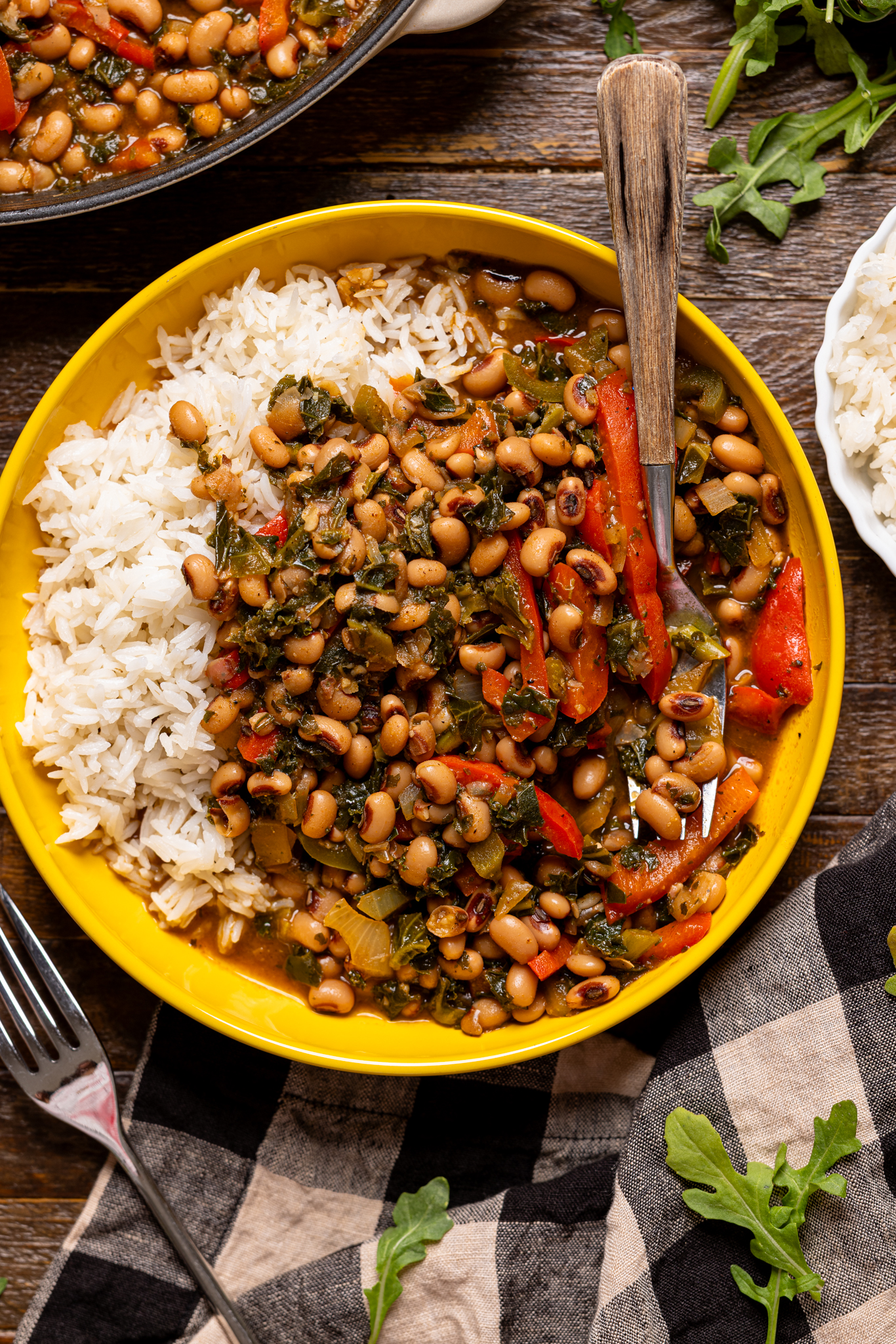 Image of cooked black eyed peas in a yellow bowl with white rice and utensils.