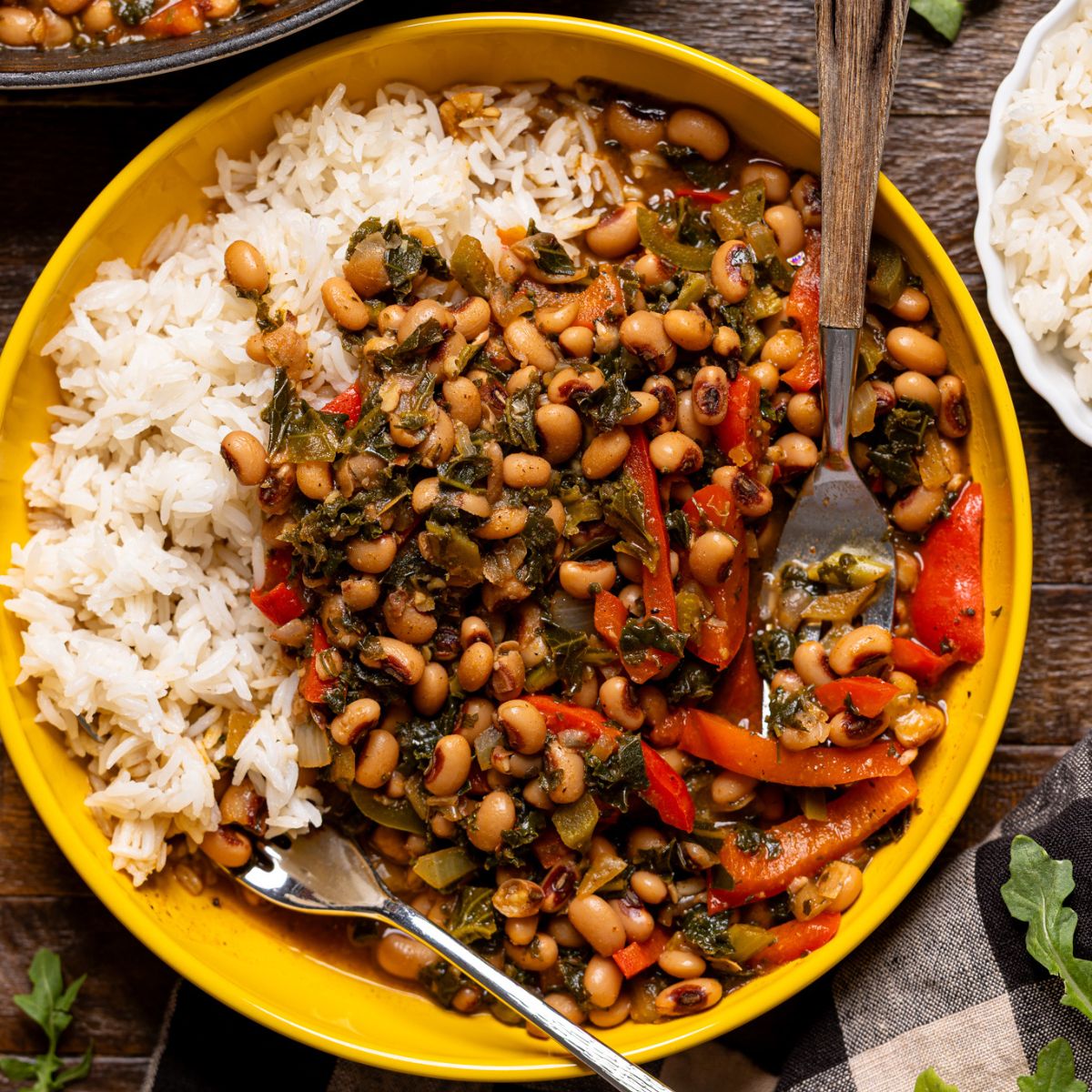 Up close shot of cooked black eyed peas in a yellow bowl with rice and utensils on a wood table.