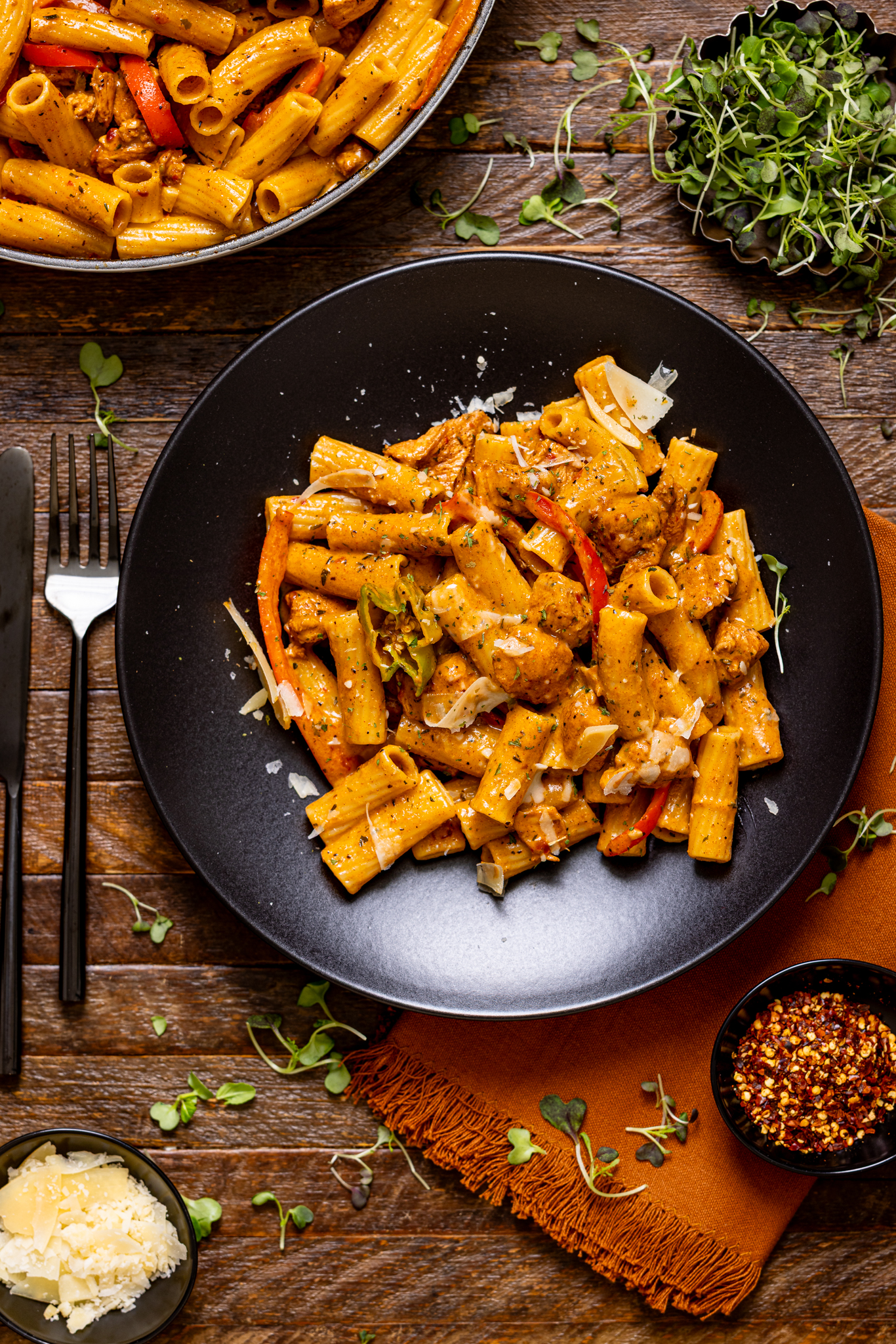Jerk chicken rasta pasta on two black plates with a side of grated parmesan. 