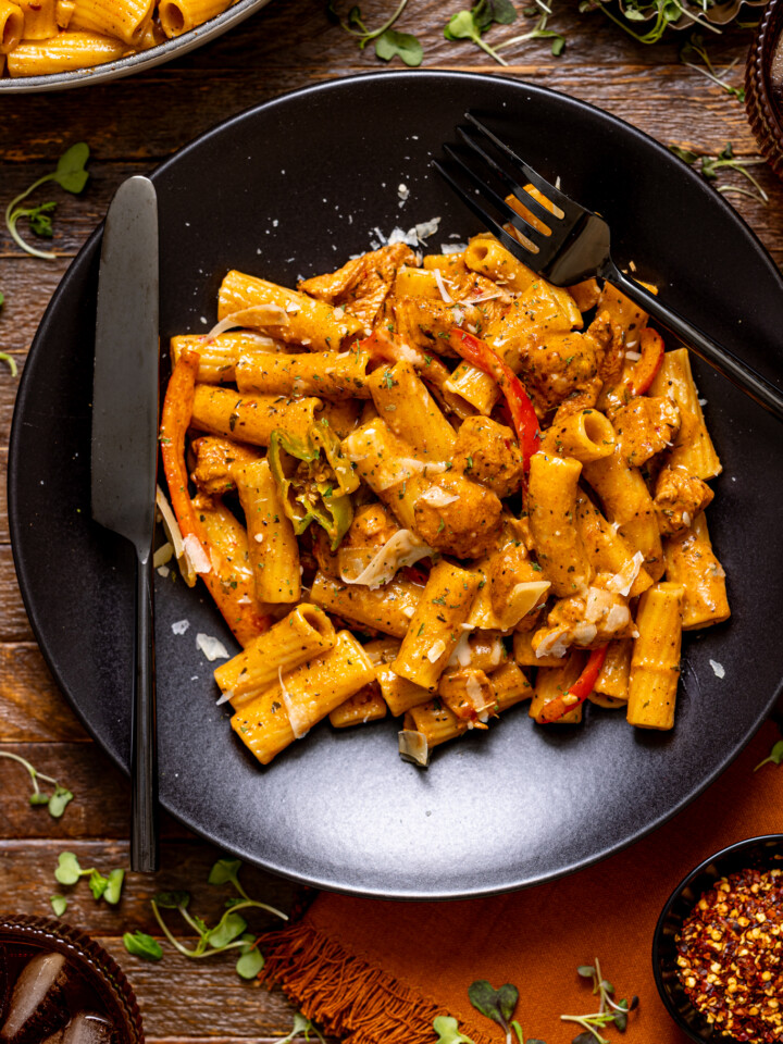 Authentic Jerk Chicken Rasta Pasta on a black plate with utensils, drinks, and red pepper flakes as garnish.