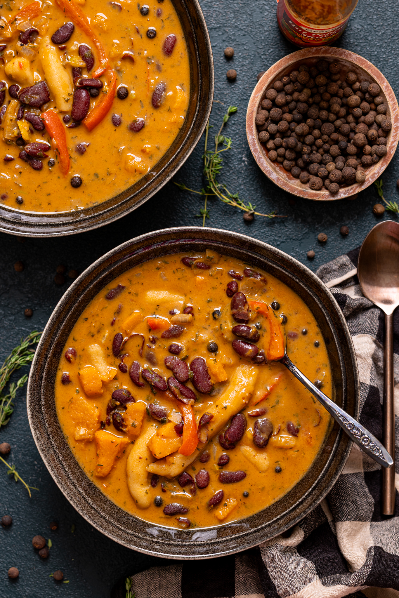 Two bowls of soup with spoons, allspice berries, and hot sauce.
