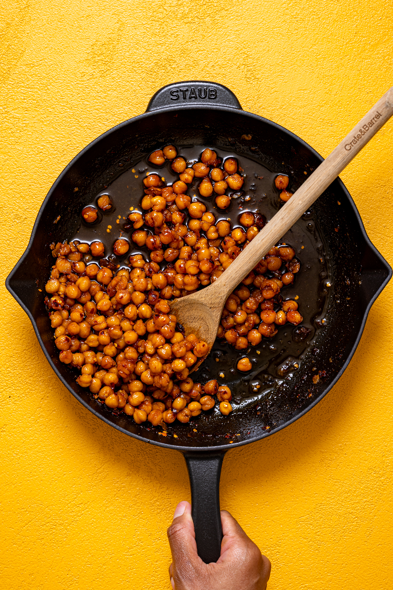 Hot honey chickpeas in a black skillet with a wooden spoon being held by woman hands.