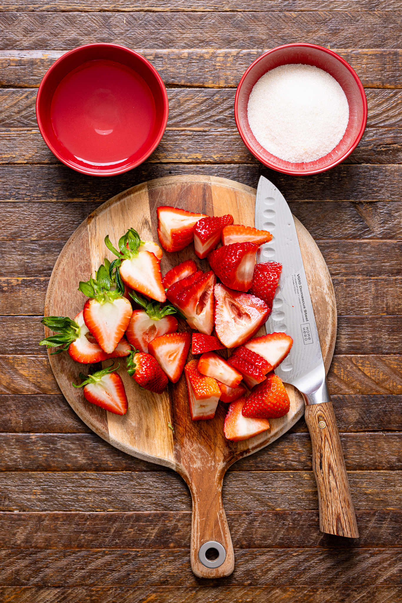 Sliced strawberries on a cutting board with a knife and ingredients on a wood table.