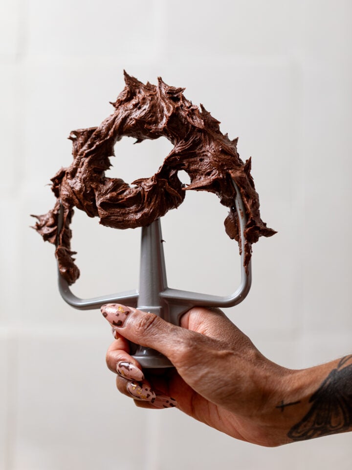 Easy Whipped Chocolate Buttercream Frosting on a mixer whisk being held by a woman's hand.