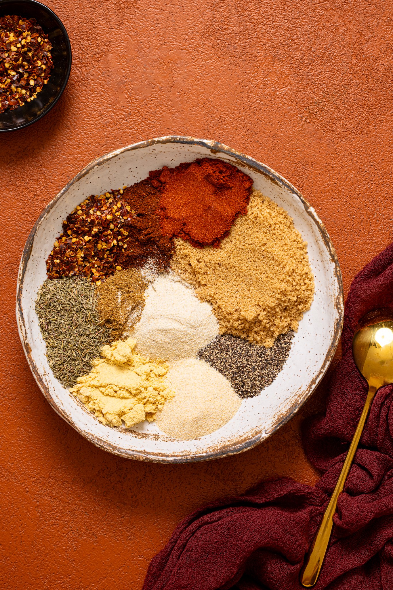 Herbs and spices in a bowl on a burnt orange table with a spoon.