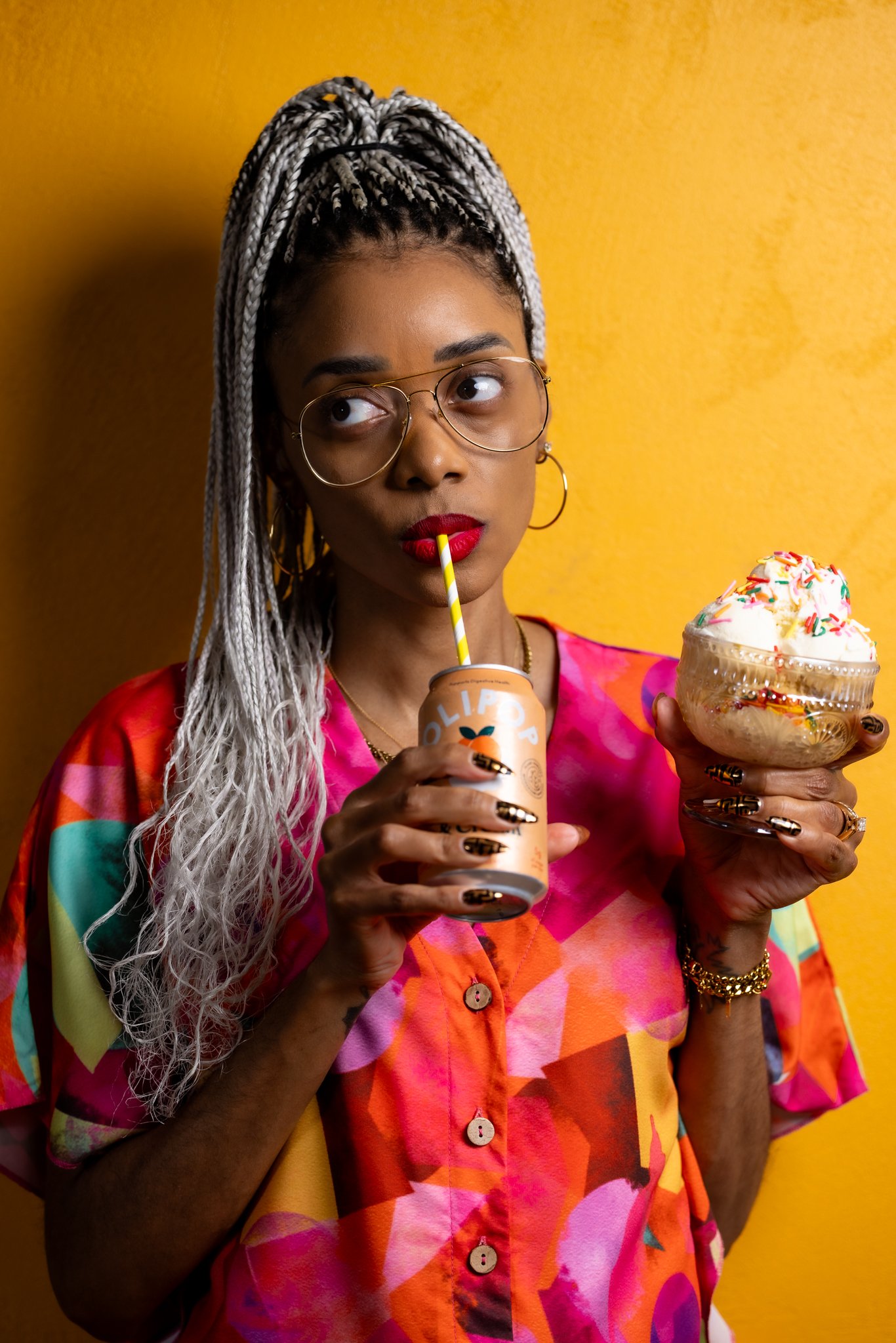 Image of Shanika in a colorful shirt with ice cream in one hand and a canned drink in another with a yellow background.