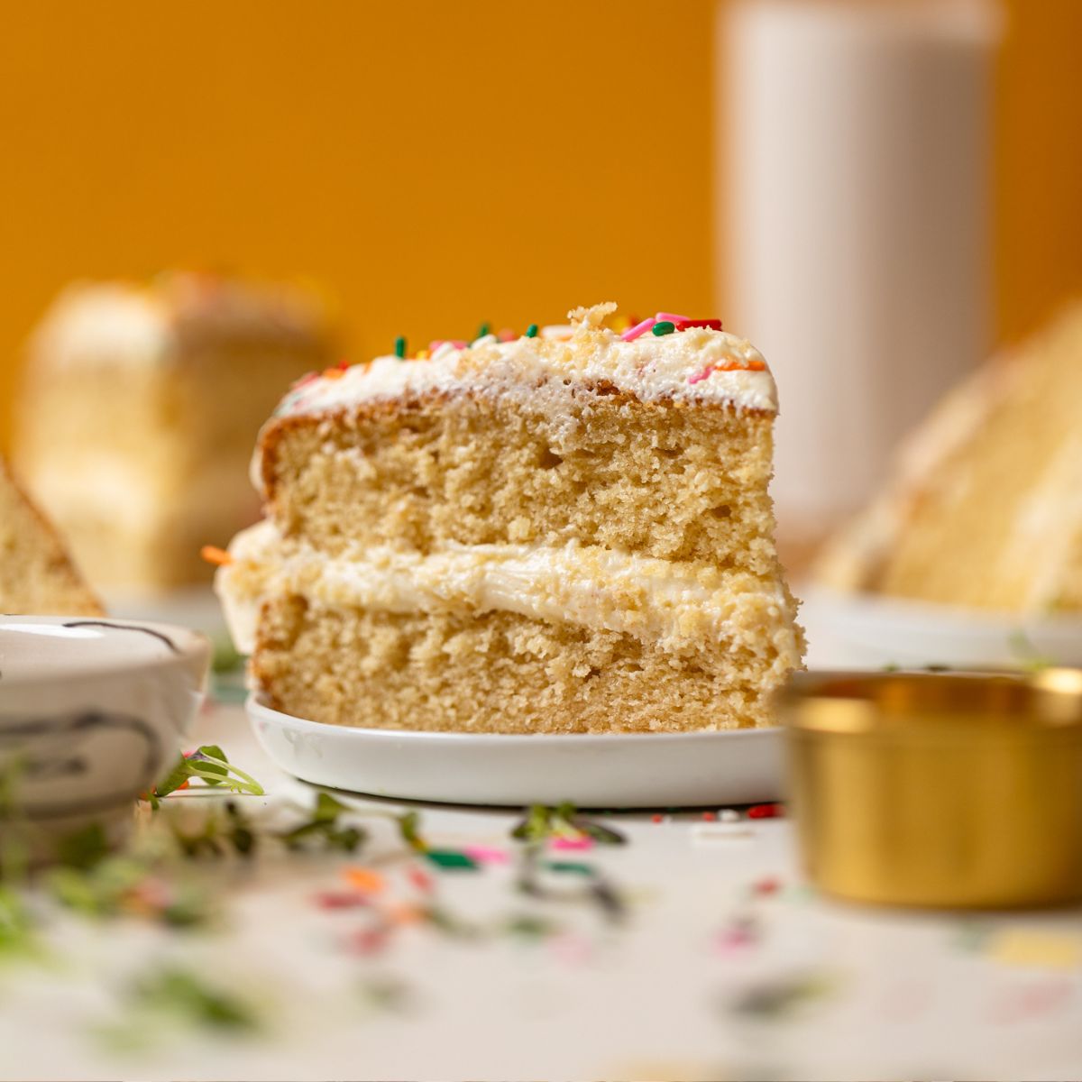 A slice of My Fave Vanilla Birthday Cake on a plate with a glass of milk in the background.