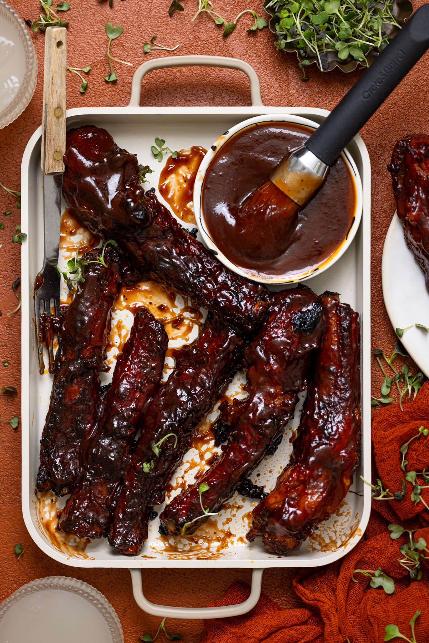 Hubby’s Favorite Oven-Baked BBQ Ribs on a baking sheet with a drink and brush with side sauce.