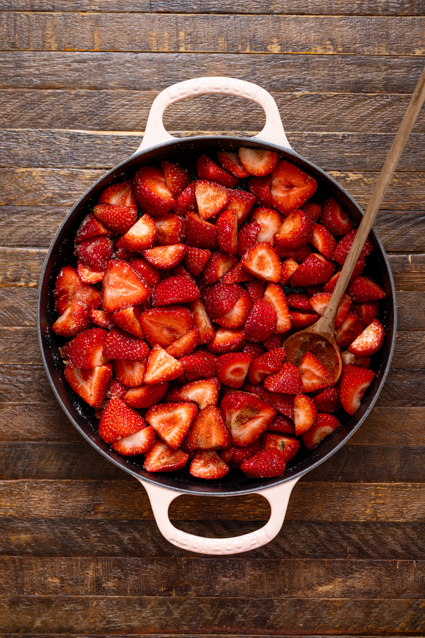 Chopped strawberries assembled in a round deep skillet with a wooden spoon.