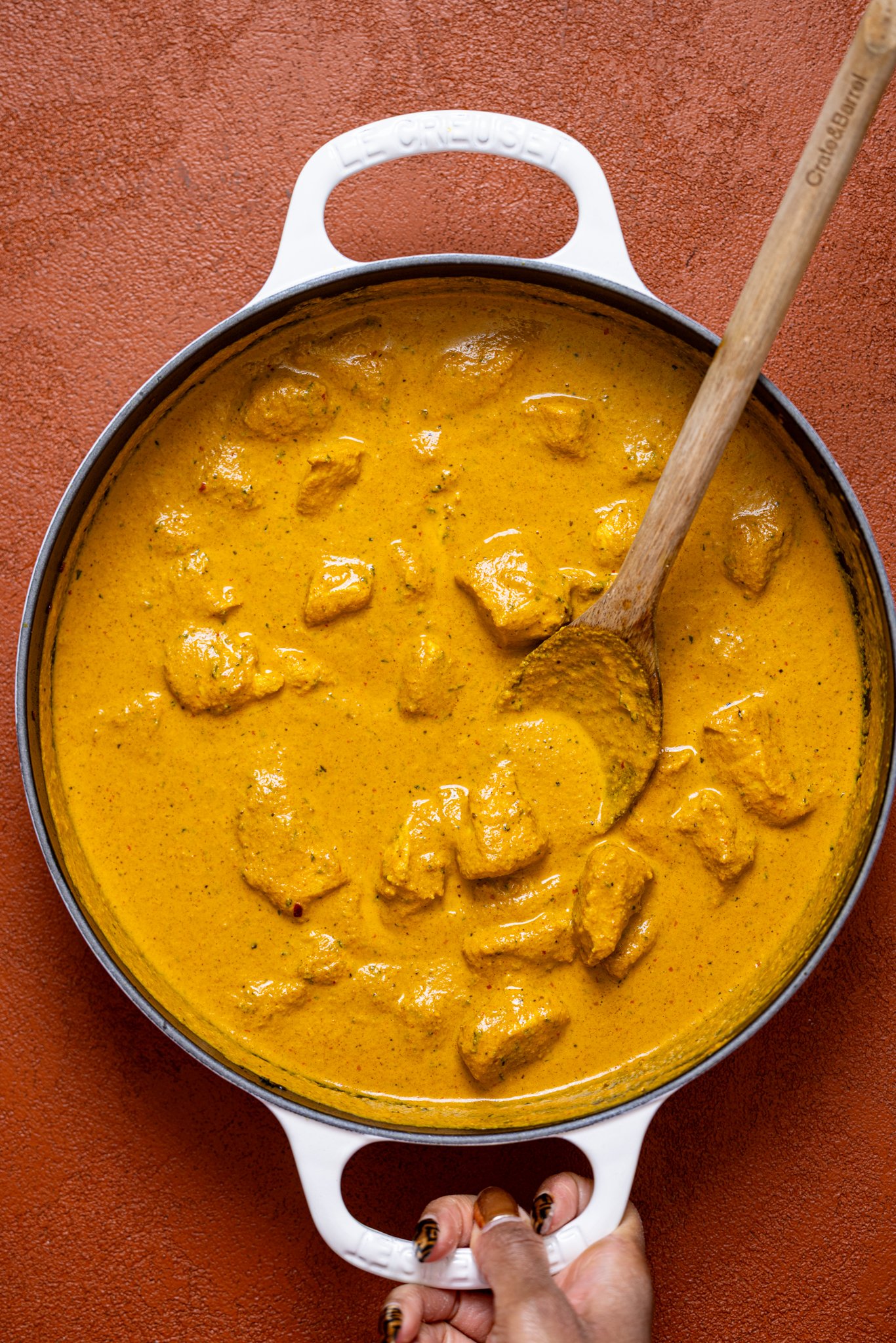 Spicy Butter Chicken in a skillet with a wooden spoon being held by woman hands.
