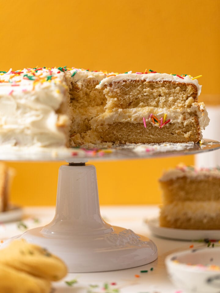 My Fave Vanilla Birthday Cake on a cake stand with a slice cut onto a plate.