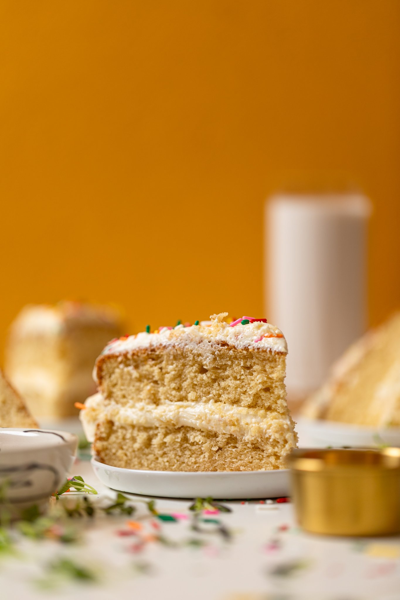 A slice of My Fave Vanilla Birthday Cake on a plate with a glass of milk in the background.